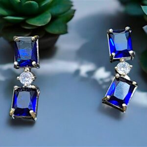 Pair of elegant blue gemstone drop earrings with rectangular blue crystals and a sparkling central stone, set against a backdrop of green succulents.