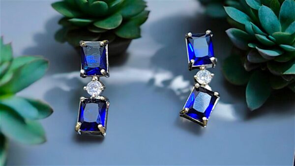 Pair of elegant blue gemstone drop earrings with rectangular blue crystals and a sparkling central stone, set against a backdrop of green succulents.