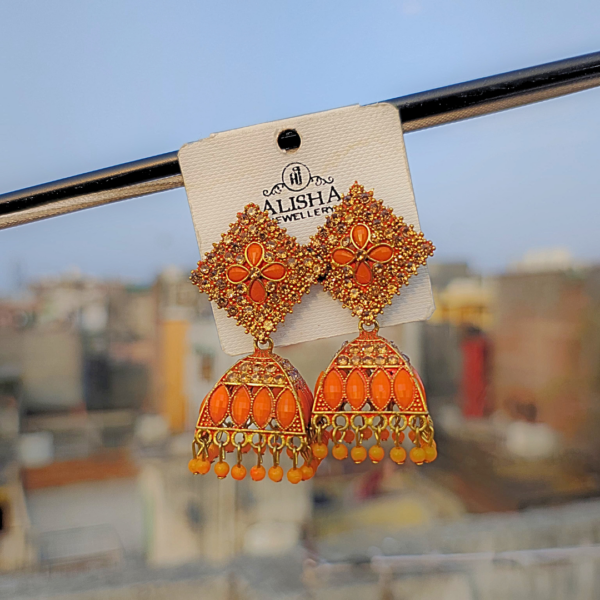 Elegant orange jhumka earrings with intricate detailing, blending traditional and trendy designs, displayed on a jewelry card against a blurred outdoor background.