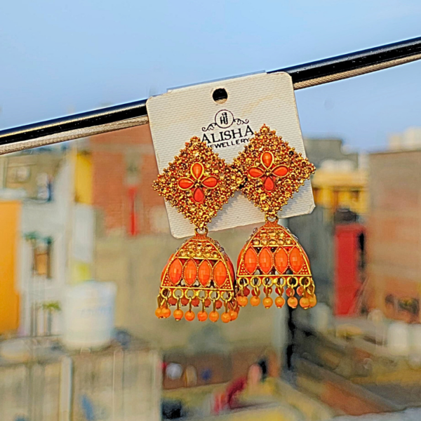 Elegant orange jhumka earrings with intricate detailing, blending traditional and trendy designs, displayed on a jewelry card against a blurred outdoor background.