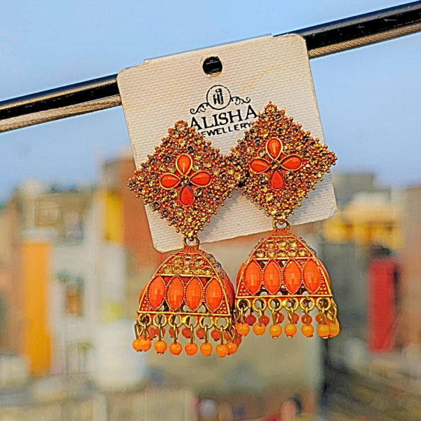 Elegant orange jhumka earrings with intricate detailing, blending traditional and trendy designs, displayed on a jewelry card against a blurred outdoor background.