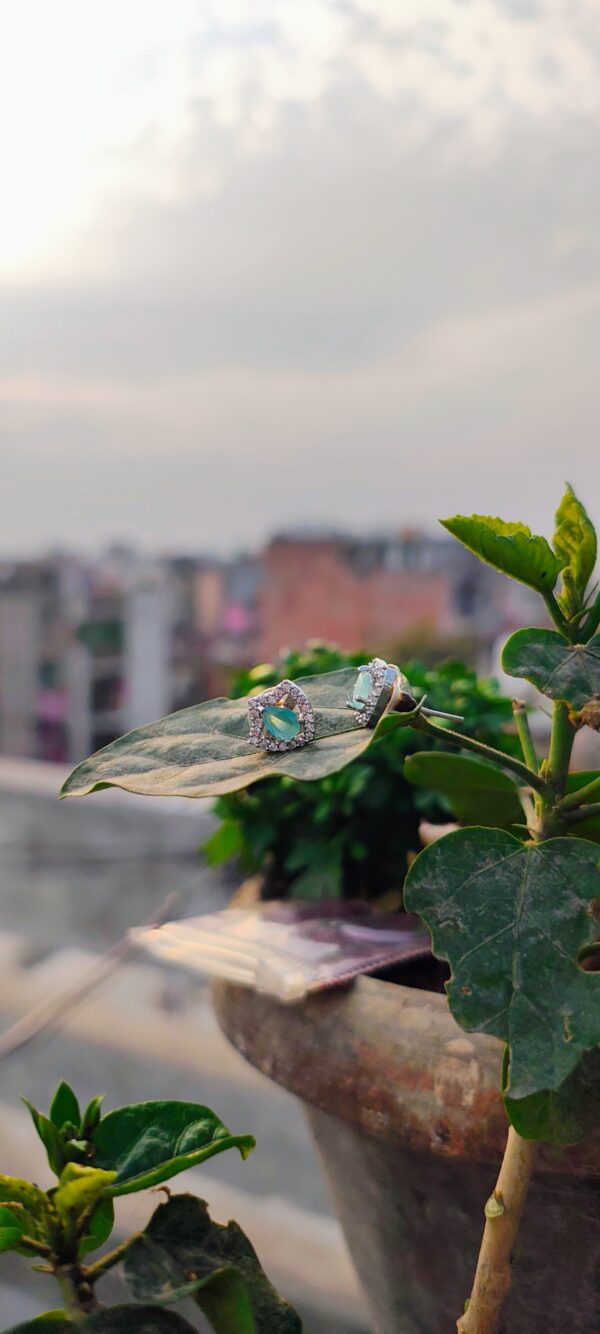 A pair of teardrop-shaped aqua stone stud earrings, encased in a sparkling crystal frame, placed on a white surface with a blurred natural background.