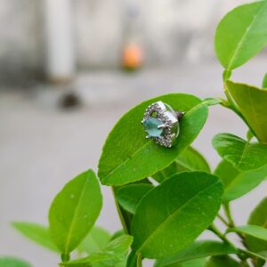 A pair of teardrop-shaped aqua stone stud earrings, encased in a sparkling crystal frame, placed on a white surface with a blurred natural background.