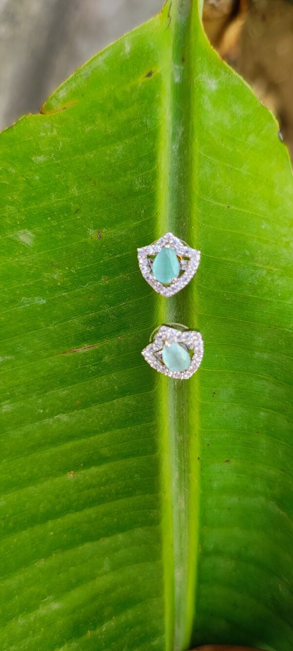 A pair of teardrop-shaped aqua stone stud earrings, encased in a sparkling crystal frame, placed on a white surface with a blurred natural background.