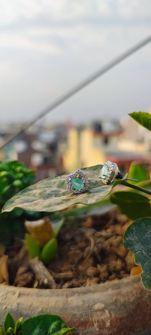 A pair of teardrop-shaped aqua stone stud earrings, encased in a sparkling crystal frame, placed on a white surface with a blurred natural background.